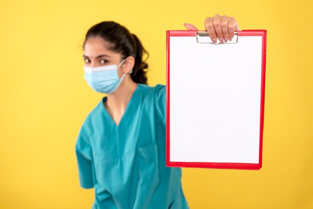 Vista frontal de la joven doctora con máscara médica sosteniendo el portapapeles en la pared amarilla