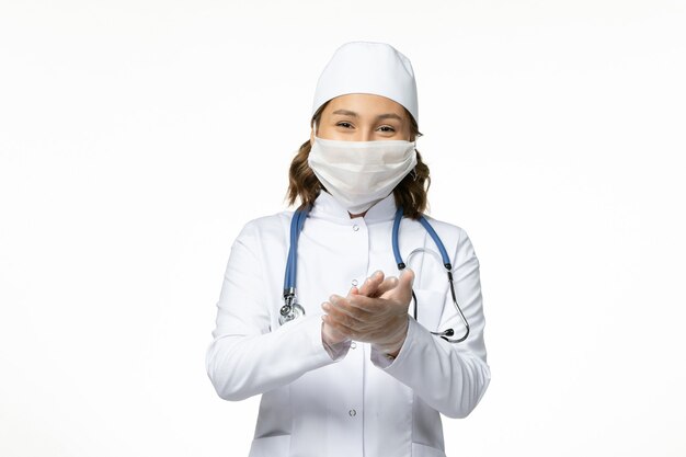 Vista frontal joven doctora con máscara protectora debido al coronavirus y sonriendo sobre una superficie blanca