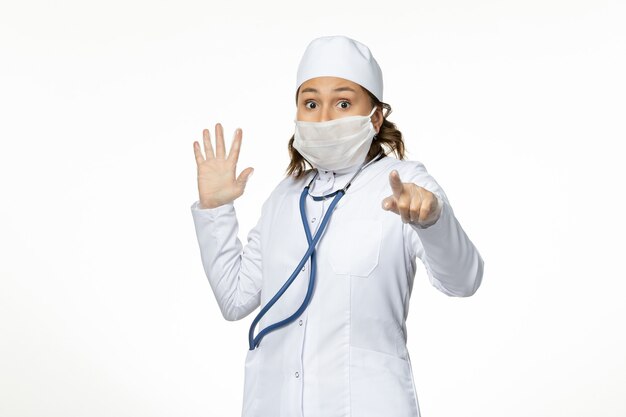 Vista frontal joven doctora con máscara protectora estéril debido al coronavirus asustada en la superficie blanca