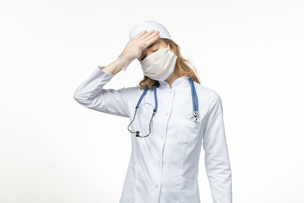 Vista frontal joven doctora con máscara protectora estéril debido al coronavirus en la superficie blanca