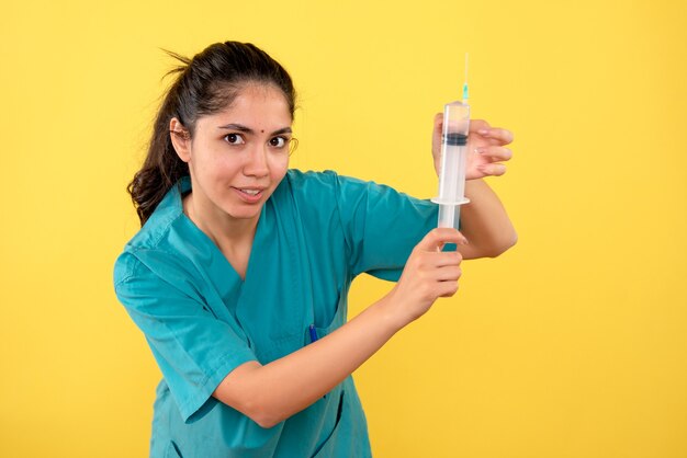 Vista frontal de la joven doctora mostrando la jeringa en la pared amarilla