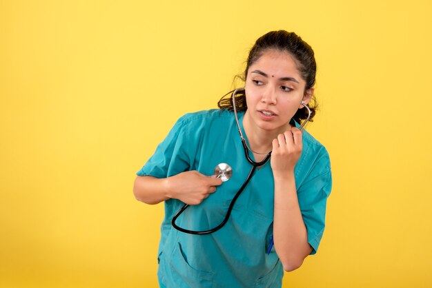 Vista frontal de la joven doctora poniendo estetoscopio en su corazón en la pared amarilla