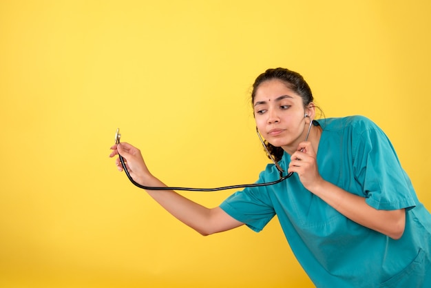 Vista frontal de la joven doctora sosteniendo un estetoscopio en la pared amarilla