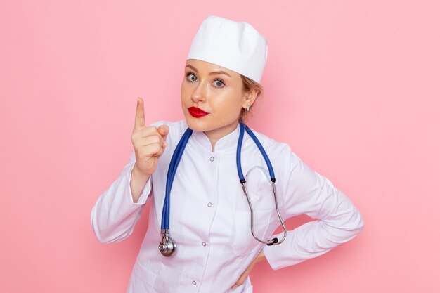 Vista frontal joven doctora en traje blanco con estetoscopio azul posando y amenazando en el trabajo femenino espacio rosa