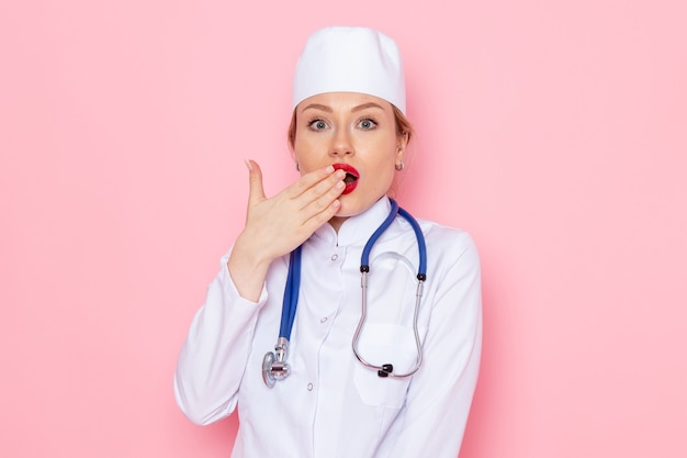 Vista frontal joven doctora en traje blanco con estetoscopio azul posando en la emoción de mujer de trabajo de espacio rosa