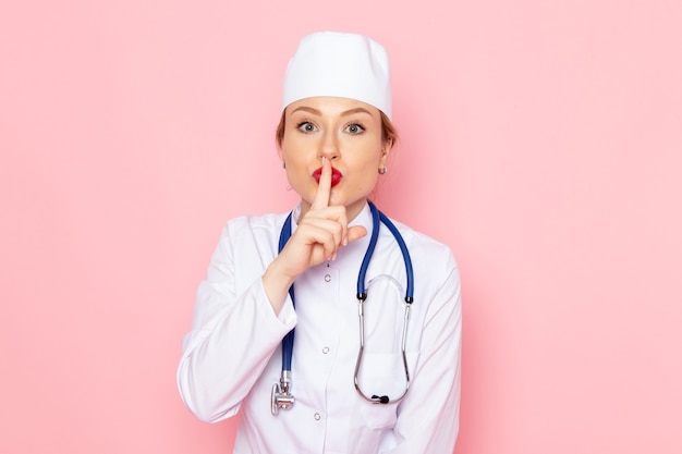 Vista frontal joven doctora en traje blanco con estetoscopio azul posando en la emoción de mujer de trabajo de espacio rosa