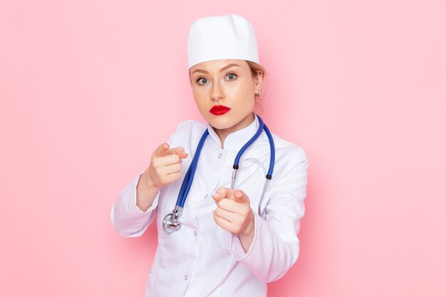 Vista frontal joven doctora en traje blanco con estetoscopio azul posando en la emoción de niña de trabajo espacial rosa