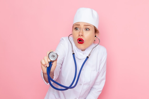 Vista frontal joven doctora en traje blanco con estetoscopio azul posando y midiendo en el espacio rosa trabajo femenino