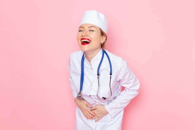 Vista frontal joven doctora en traje blanco con estetoscopio azul posando y riendo en la emoción de mujer de trabajo de espacio rosa