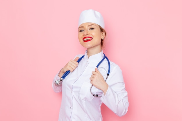 Vista frontal joven doctora en traje blanco con estetoscopio azul posando sonriendo en el espacio rosa trabajo medicina médico hospital médico