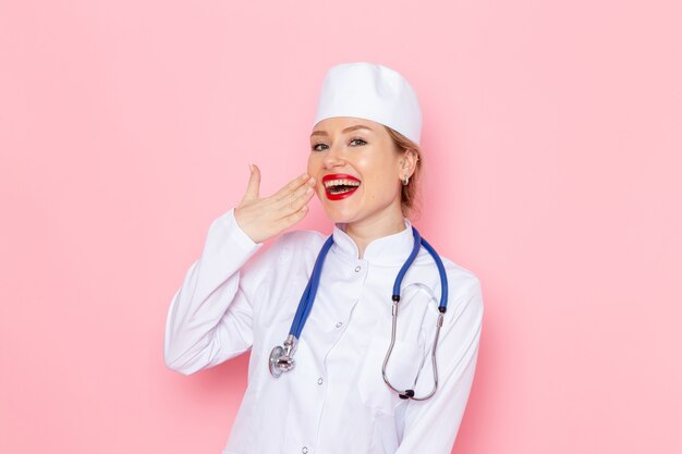 Vista frontal joven doctora en traje blanco con estetoscopio azul posando y sonriendo en el trabajo espacial rosa