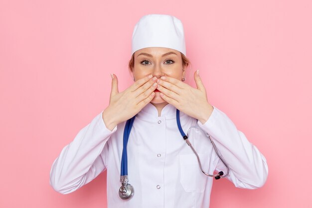 Vista frontal joven doctora en traje blanco con estetoscopio azul posando y sonriendo en el trabajo femenino del hospital médico de medicina espacial rosa