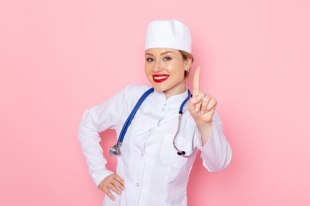 Vista frontal joven doctora en traje blanco con estetoscopio azul sonriendo y posando en el hospital médico de medicina espacial rosa femenino