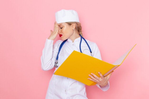 Vista frontal joven doctora en traje blanco con estetoscopio azul sosteniendo archivos amarillos en el espacio rosa femenino
