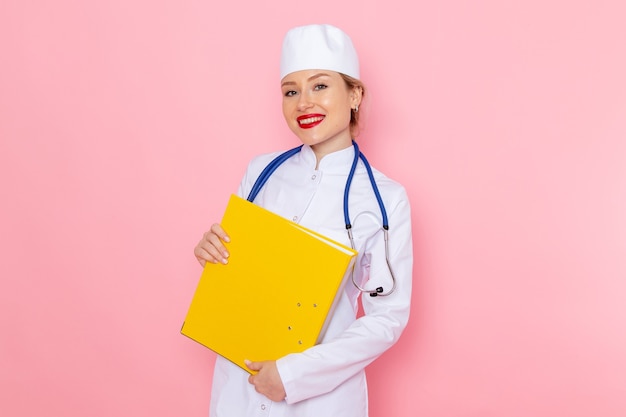 Vista frontal joven doctora en traje blanco con estetoscopio azul sosteniendo archivos amarillos en el espacio rosa trabajo femenino