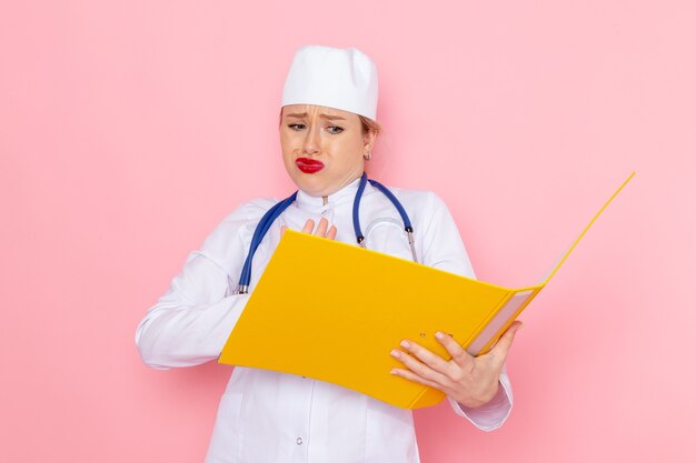 Vista frontal joven doctora en traje blanco con estetoscopio azul sosteniendo archivos amarillos en el trabajo femenino del hospital médico de medicina espacial rosa