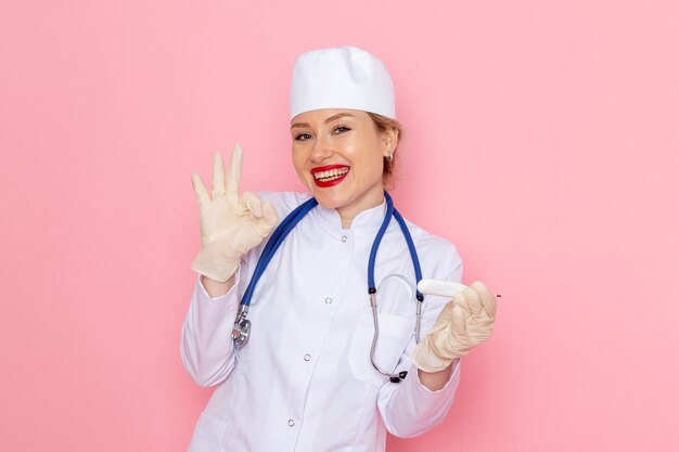 Vista frontal joven doctora en traje médico blanco con dispositivo de sujeción de estetoscopio azul sonriendo en el hospital médico de medicina espacial rosa