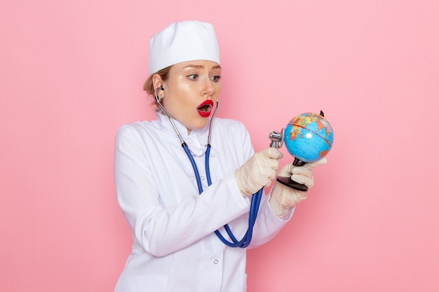 Vista frontal joven doctora en traje médico blanco con estetoscopio azul comprobando el pequeño globo en el trabajo de hospital médico de medicina espacial rosa