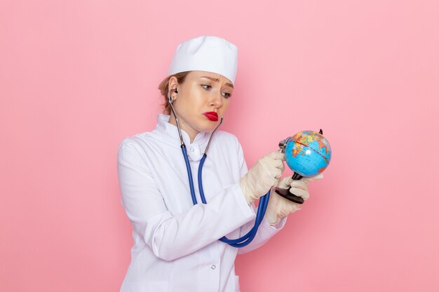 Vista frontal joven doctora en traje médico blanco con estetoscopio azul comprobando el pequeño globo en el trabajo del hospital médico de medicina espacial rosa