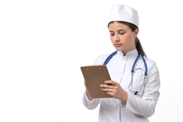 Una vista frontal joven doctora en traje médico blanco con estetoscopio azul escribiendo notas