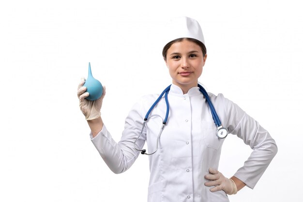 Una vista frontal joven doctora en traje médico blanco con estetoscopio azul y guantes con enema