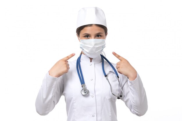 Una vista frontal joven doctora en traje médico blanco con estetoscopio azul con máscara blanca