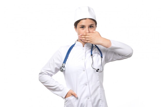 Una vista frontal joven doctora en traje médico blanco con estetoscopio azul posando con expresión tímida