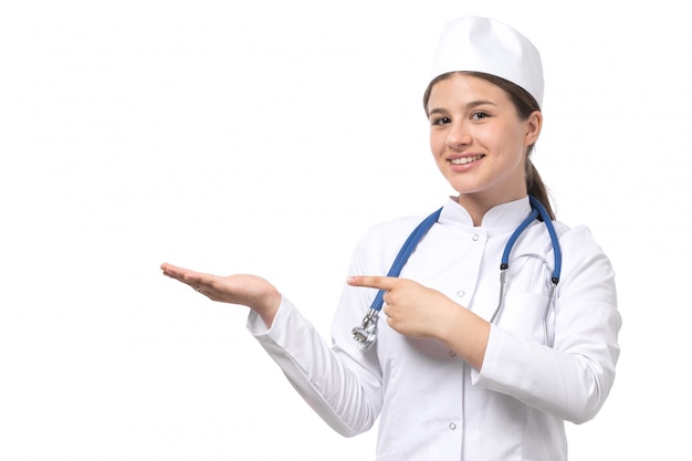 Una vista frontal joven doctora en traje médico blanco con estetoscopio azul posando con sonrisa