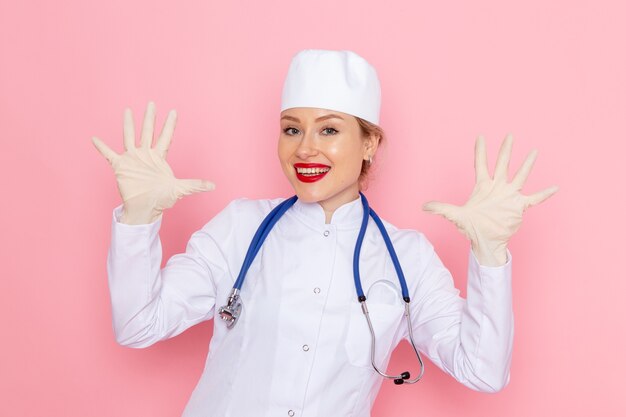 Vista frontal joven doctora en traje médico blanco con estetoscopio azul sonriendo con guantes en el trabajador del hospital médico de medicina espacial rosa