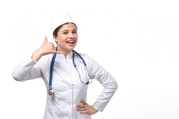 Una vista frontal joven doctora en traje médico blanco con estetoscopio azul sonriendo y posando