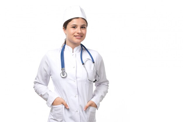 Una vista frontal joven doctora en traje médico blanco con estetoscopio azul sonriendo