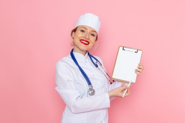 Vista frontal joven doctora en traje médico blanco con estetoscopio azul sosteniendo el bloc de notas con una sonrisa en el hospital médico de medicina espacial rosa