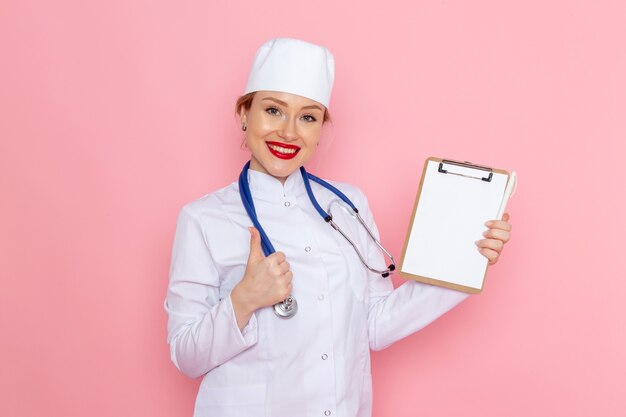 Vista frontal joven doctora en traje médico blanco con estetoscopio azul sosteniendo el bloc de notas con una sonrisa en la salud del hospital médico de medicina espacial rosa