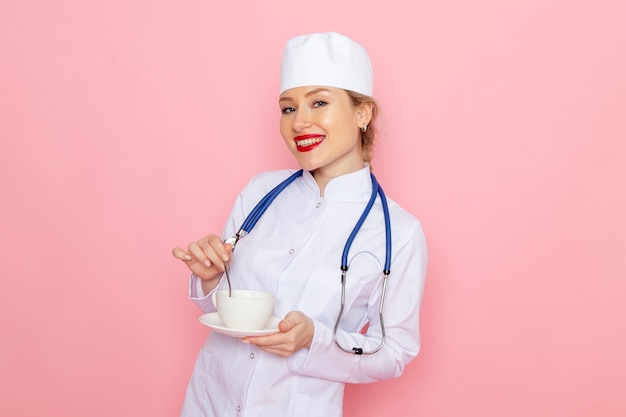 Vista frontal joven doctora en traje médico blanco con estetoscopio azul sosteniendo café sonriendo en el hospital médico de medicina espacial rosa