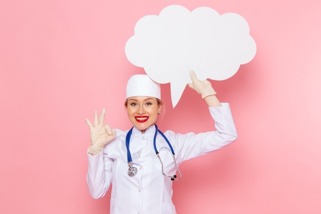 Vista frontal joven doctora en traje médico blanco con estetoscopio azul sosteniendo un enorme cartel blanco y sonriendo en la salud del hospital médico de medicina espacial rosa