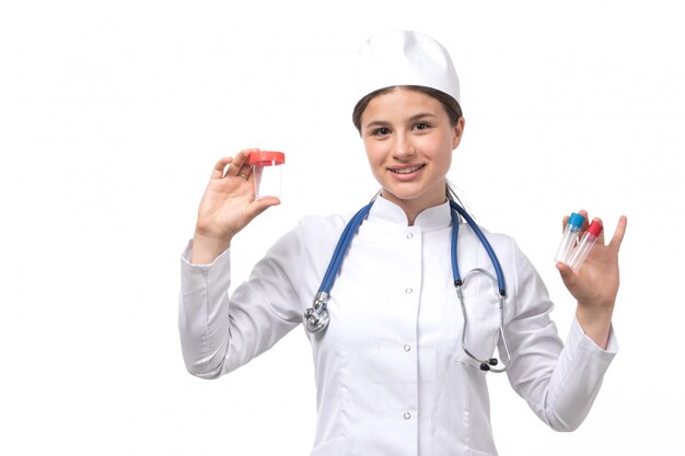 Una vista frontal joven doctora en traje médico blanco con estetoscopio azul sosteniendo frascos y sonriendo