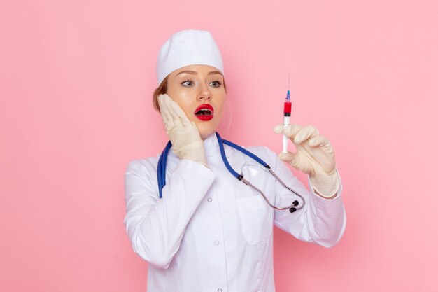 Vista frontal joven doctora en traje médico blanco con estetoscopio azul sosteniendo la inyección en la mujer de trabajador de hospital médico de medicina espacial rosa