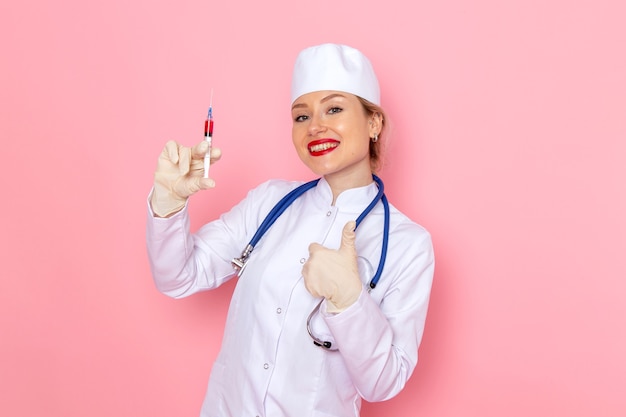 Vista frontal joven doctora en traje médico blanco con estetoscopio azul sosteniendo la inyección con una sonrisa en la mujer de trabajador de hospital médico de medicina espacial rosa