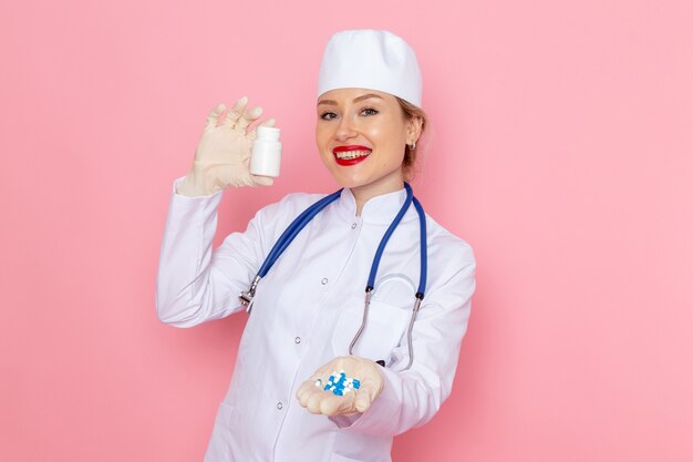 Vista frontal joven doctora en traje médico blanco con estetoscopio azul sosteniendo pastillas con sonrisa en el hospital médico de medicina espacial rosa