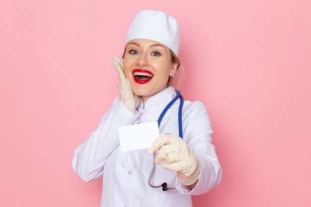 Vista frontal joven doctora en traje médico blanco con estetoscopio azul sosteniendo una tarjeta blanca sonriendo en la enfermera del hospital médico de medicina espacial rosa