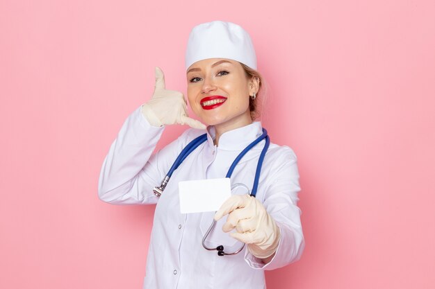 Vista frontal joven doctora en traje médico blanco con estetoscopio azul sosteniendo una tarjeta blanca sonriendo posando en el espacio rosa