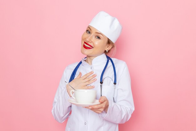 Vista frontal joven doctora en traje médico blanco con estetoscopio azul sosteniendo una taza de café con una sonrisa en el hospital médico de medicina espacial rosa