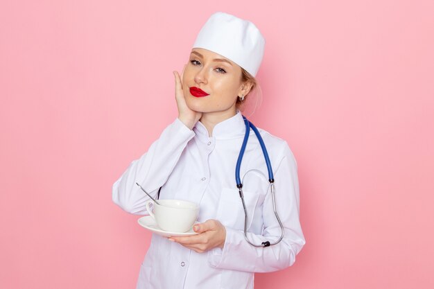 Vista frontal joven doctora en traje médico blanco con estetoscopio azul sosteniendo una taza de café en el trabajo de hospital médico de medicina espacial rosa