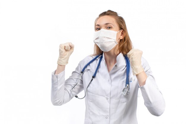 Una vista frontal joven doctora en traje médico blanco con estetoscopio con máscara protectora blanca en guantes en el blanco