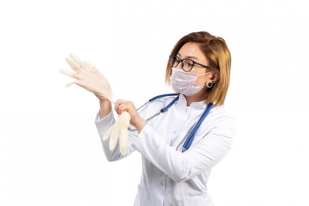 Una vista frontal joven doctora en traje médico blanco con estetoscopio con máscara protectora blanca con guantes en el blanco