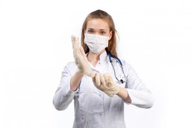 Foto gratuita una vista frontal joven doctora en traje médico blanco con estetoscopio con máscara protectora blanca con guantes en el blanco