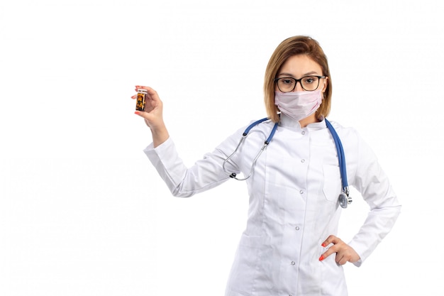 Una vista frontal joven doctora en traje médico blanco con estetoscopio con máscara protectora blanca con pastillas en el blanco