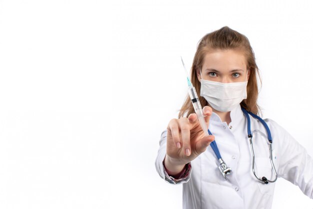 Una vista frontal joven doctora en traje médico blanco con estetoscopio con máscara protectora blanca posando con inyección en el blanco