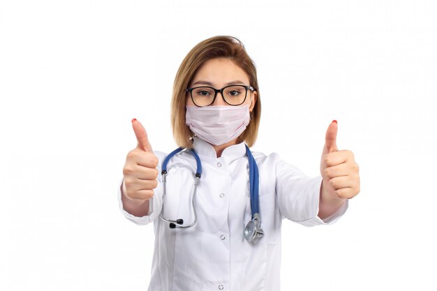 Una vista frontal joven doctora en traje médico blanco con estetoscopio con máscara protectora blanca posando mostrando como signo en el blanco