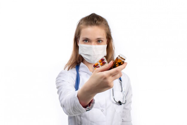 Una vista frontal joven doctora en traje médico blanco con estetoscopio con máscara protectora blanca posando sosteniendo pastillas en el blanco